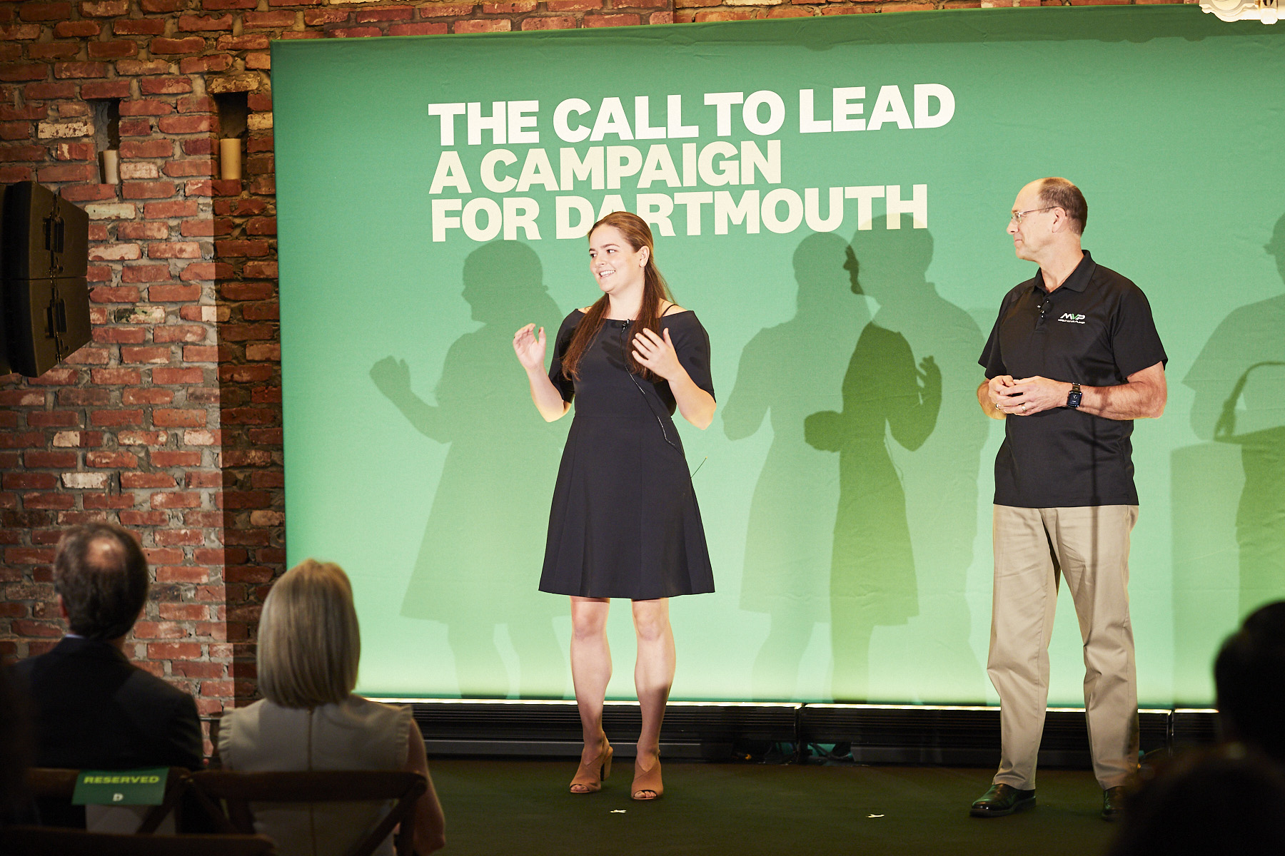 A student and professor on stage in front of a large green backdrop.