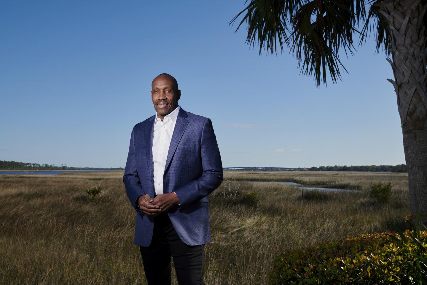 A photo of a man in a blue blazer with hands folded in front of him in an outdoor landscape