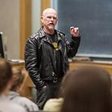 A journalist wearing a leather jacket and goatee speaks at Dartmouth