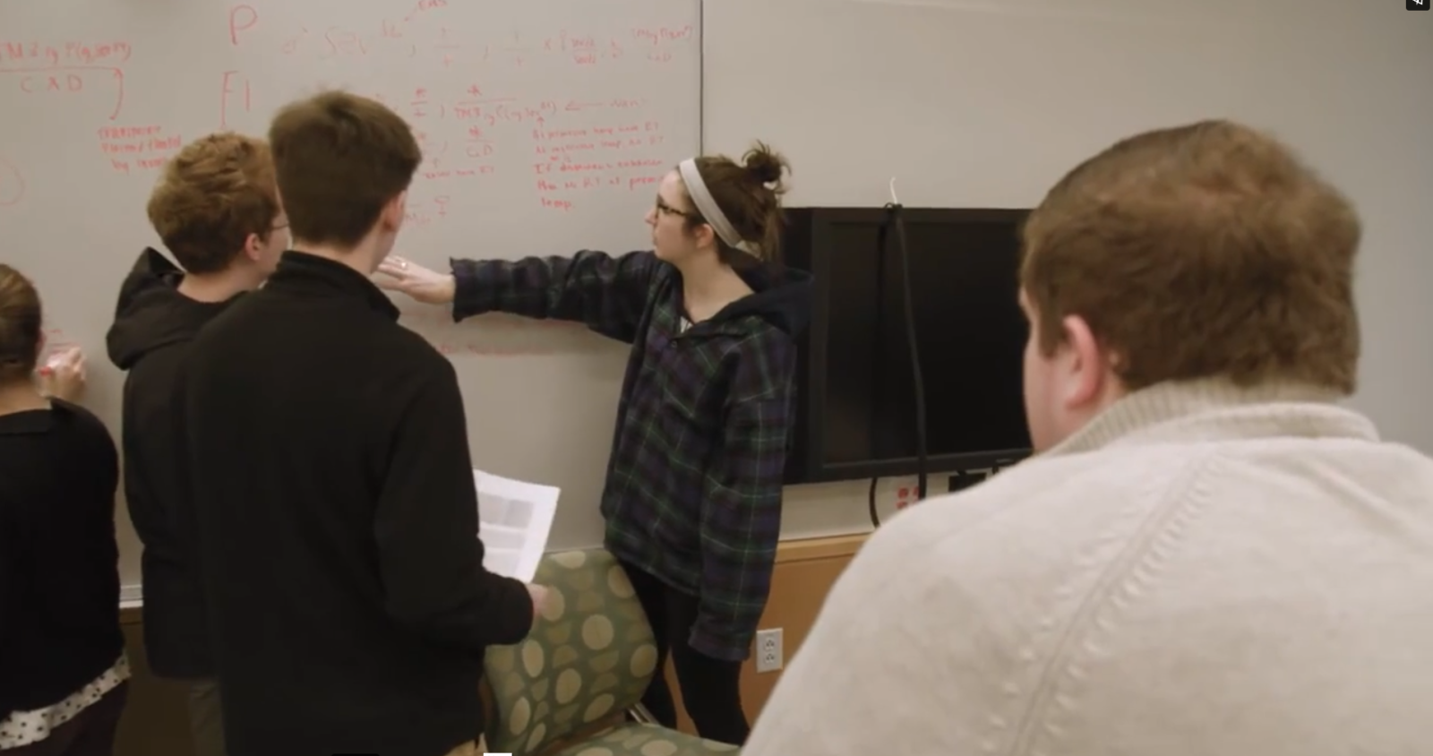 one student speaking to a group of other students while pointing to a white board