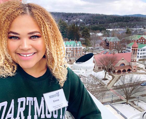 A smiling woman stands on a tower balcony, with the campus below.